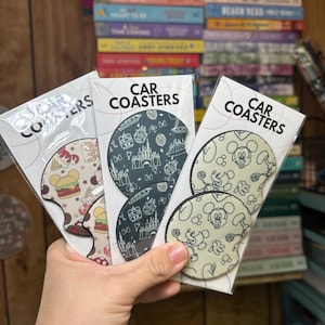 May include: Three sets of car coasters featuring different Disney designs. The first set has a black and white design with a castle, fireworks, and other Disney icons. The second set has a black and white design with Mickey Mouse and other Disney characters. The third set has a red, white, and blue design with a hamburger, ice cream cone, and other Disney food items.