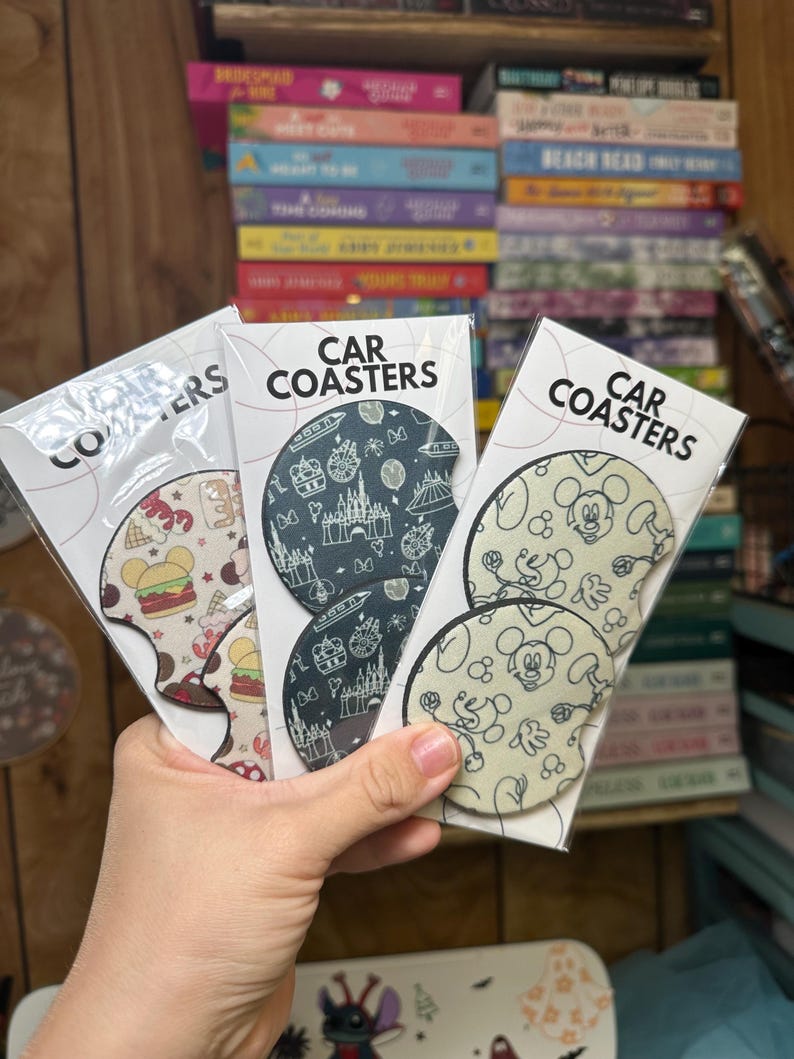 May include: Three sets of car coasters featuring different Disney designs. The first set has a black and white design with a castle, fireworks, and other Disney icons. The second set has a black and white design with Mickey Mouse and other Disney characters. The third set has a red, white, and blue design with a hamburger, ice cream cone, and other Disney food items.