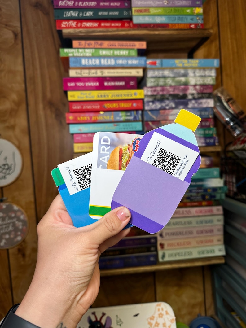 May include: A hand holds three gift cards. The cards are blue, purple, and white. The blue card has a QR code and the text "To Connect!" The purple card has a QR code and the text "To Connect!" The white card has a picture of a hamburger and the text "CARD".