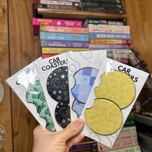 May include: A set of six car coasters featuring different patterns. The coasters are made of a durable material and are designed to protect your car's cup holders from spills and scratches. The patterns include a blue plaid, a yellow floral design, a green butterfly design, and a black and white star design. The coasters are packaged in clear plastic sleeves and labeled "CAR COASTERS".