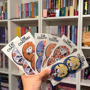 May include: A collection of circular car coasters packaged in clear plastic sleeves, each featuring Snoopy in various Halloween-themed illustrations including pumpkins, witches, and autumn leaves, with the text Car Coasters printed on each label.