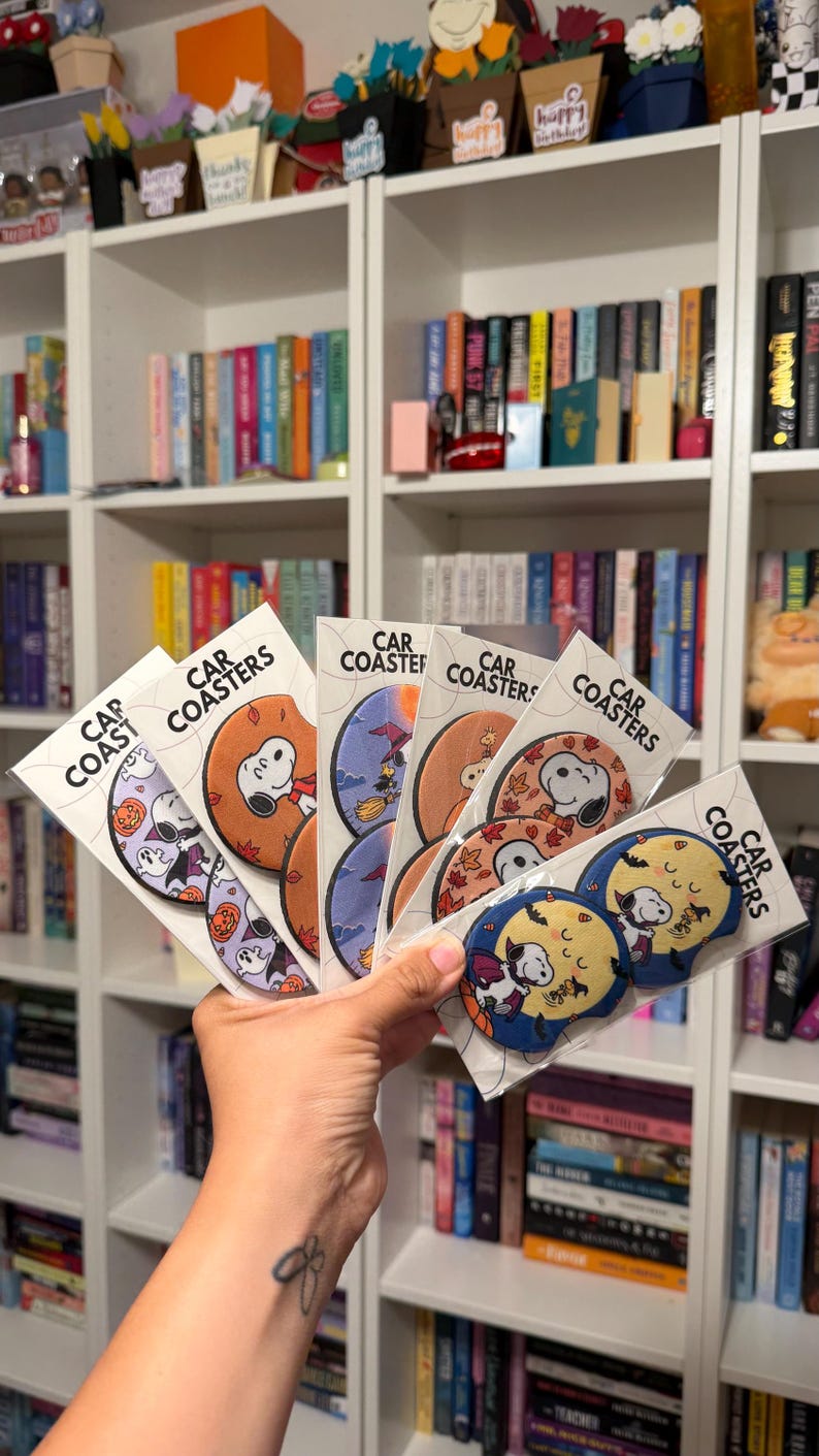 May include: A collection of circular car coasters packaged in clear plastic sleeves, each featuring Snoopy in various Halloween-themed illustrations including pumpkins, witches, and autumn leaves, with the text Car Coasters printed on each label.