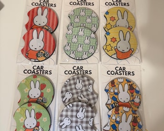 May include: Six sets of two circular car coasters in clear plastic packaging, each labeled CAR COASTERS. The coasters feature Miffy the rabbit in various patterns including red stripes, green gingham, yellow stars, strawberries, gray plaid, and multi-colored icons.