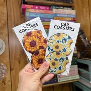 May include: Two sets of car coasters featuring a sunflower design. One set has a yellow and blue sunflower pattern on a white background. The other set has a red and yellow sunflower pattern on a black background. The packaging says "Car Coasters".