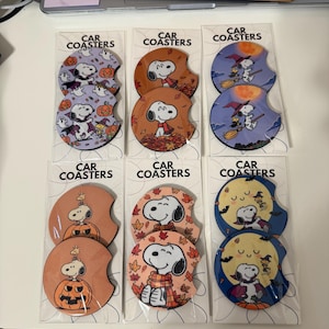May include: Six sets of two circular car coasters in clear plastic packaging, each labeled CAR COASTERS. The coasters feature Snoopy and Woodstock in various Halloween and autumn themes, including pumpkins, ghosts, witches, and falling leaves.