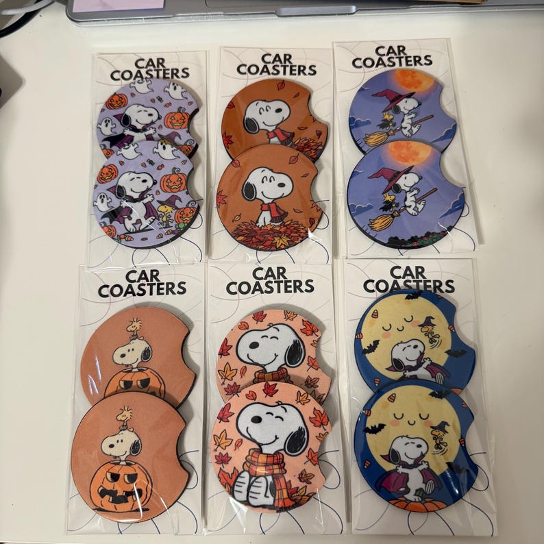 May include: Six sets of two circular car coasters in clear plastic packaging, each labeled CAR COASTERS. The coasters feature Snoopy and Woodstock in various Halloween and autumn themes, including pumpkins, ghosts, witches, and falling leaves.