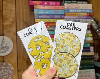 May include: Two round yellow and white car coasters with a cartoon egg character design. The coasters are packaged in clear plastic and labeled "CAR COASTERS".
