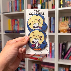 May include: Two round car coasters in a clear plastic sleeve with the text CAR COASTERS at the top. Each coaster features a graphic of a white dog dressed as a vampire standing on a pumpkin against a large yellow moon and dark blue sky.