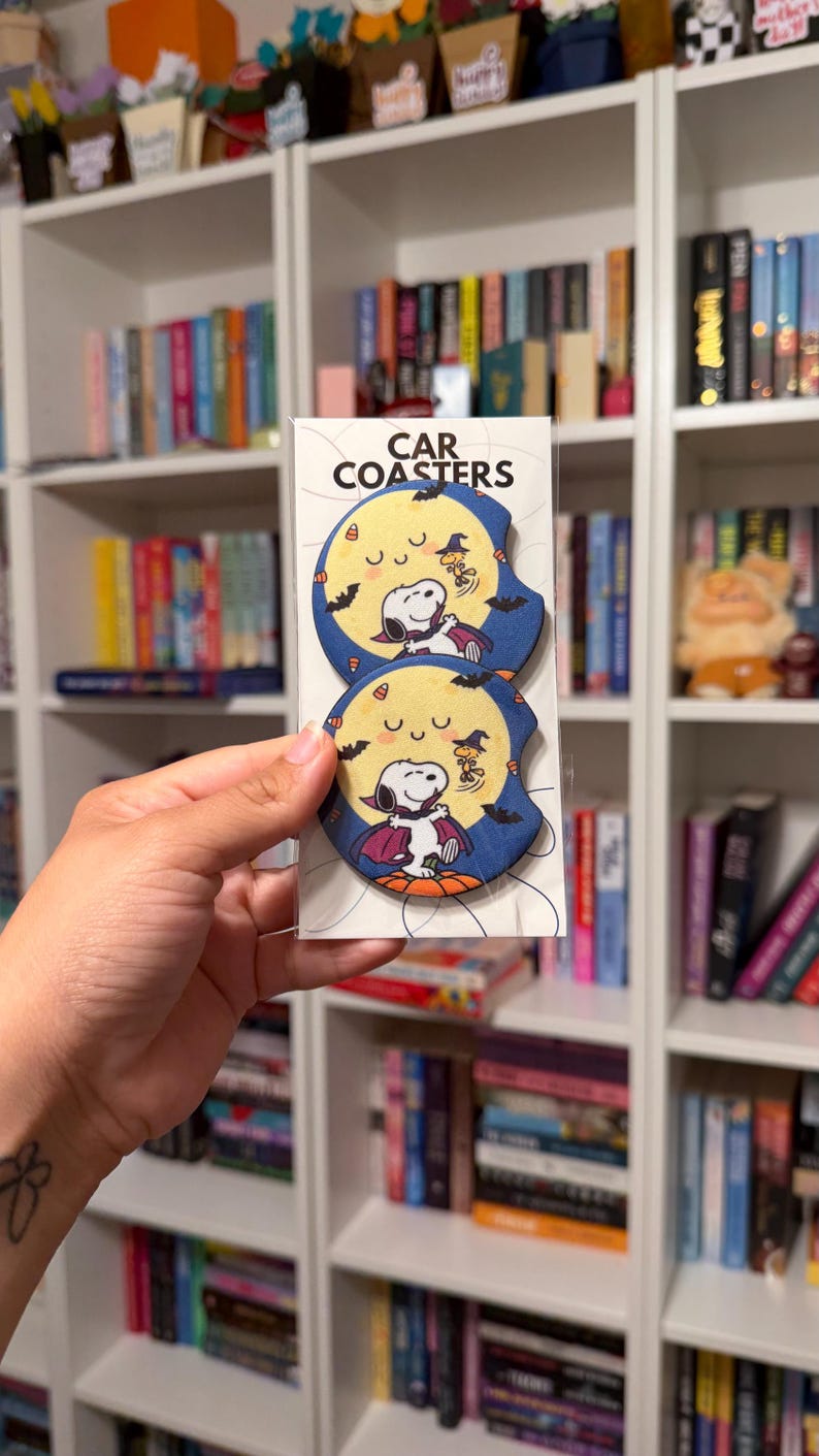 May include: Two round car coasters in a clear plastic sleeve with the text CAR COASTERS at the top. Each coaster features a graphic of a white dog dressed as a vampire standing on a pumpkin against a large yellow moon and dark blue sky.
