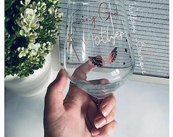 Può includere: Un bicchiere di vino in vetro trasparente con un design di glitter dorati che dice "Fairy God Mother" con una stella e ali. Il bicchiere è tenuto da una mano.