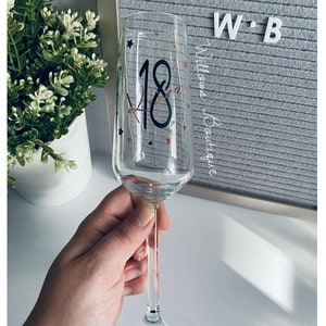 May include: A clear glass champagne flute with a black number 18 and gold glitter stars. The glass is held in a hand.