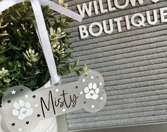May include: A bone-shaped pet tag with the name "Misty" in script. The tag is clear with white paw prints and gold stars. It hangs from a white ribbon, and is next to a white pot with greenery. The text "Willow's Boutique" is in the background.