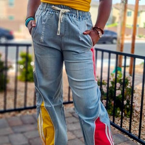 May include: Light blue denim pants with yellow and red side panels. The pants have a drawstring waist and distressed details. The side panels have white stripes. The pants are part of a casual clothing collection.