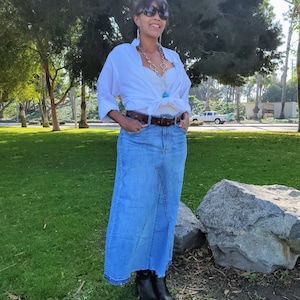 May include: A woman wearing a white button-down shirt tied at the waist, a long denim skirt, and black ankle boots. She is wearing a silver necklace with a turquoise pendant and sunglasses.