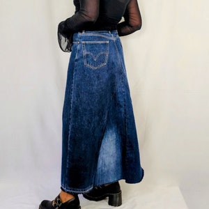 May include: A woman wearing a black sheer long-sleeved top and a dark blue denim maxi skirt. The skirt has a back slit and a classic five-pocket design.