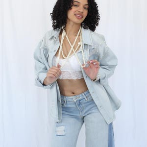 Puede incluir: Chaqueta vaquera azul claro adornada con perlas, combinada con vaqueros de lavado claro. Un bralette de encaje blanco y un largo collar de perlas completan el conjunto. Los vaqueros tienen detalles desgastados.