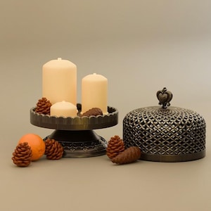 May include: A decorative arrangement featuring three cream-colored candles on a dark metal pedestal, surrounded by small pinecones and an orange. A decorative metal cloche with a crown-shaped handle sits nearby. The items are arranged on a neutral background.