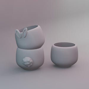 Peut inclure: Trois tasses en céramique gris clair. Deux sont empilées, ressemblant à un lapin avec des détails d'oreilles et de queue. La troisième tasse est de conception simple et arrondie. Les tasses sont probablement à usage décoratif ou fonctionnel.