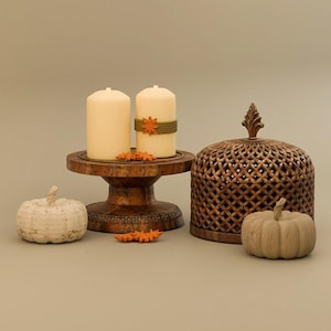 May include: A decorative fall-themed arrangement featuring two cream-colored pillar candles on a wooden pedestal, a carved wooden lantern, and two small pumpkin figurines. The candles are adorned with a green and orange decorative band.