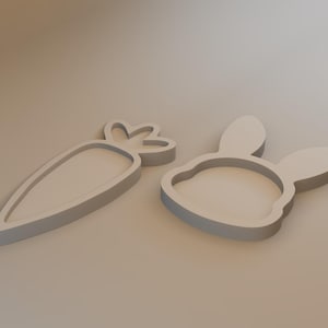 May include: Two light gray cookie cutters in the shapes of a carrot and a bunny head. The carrot cutter has a detailed top, and the bunny cutter has long ears and a rounded face. The items are likely for Easter or spring baking.