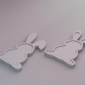 Puede incluir: Dos decoraciones en forma de conejito gris claro. Cada conejito tiene un cuerpo redondeado, orejas largas y una pequeña cola. Las decoraciones están colocadas sobre una superficie gris claro, creando una estética minimalista.