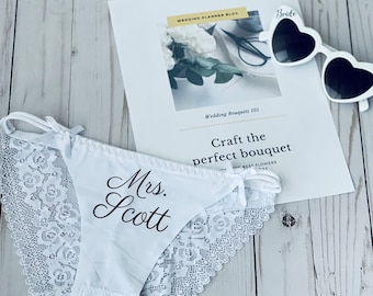 May include: White bridal lingerie with lace trim and the name "Mrs. Scott" printed in black script. A pair of heart-shaped sunglasses with "Bride" on the frame and a wedding planning blog are also in the image.