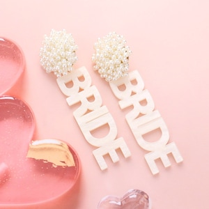 May include: A pair of white 'BRIDE' earrings with pearl embellishments. The earrings are on a pink background.