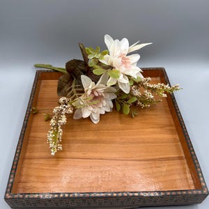 Könnte beinhalten: Ein quadratisches Holztablett mit dekorativem Rand, das einen Strauß weißer und rosa Blumen mit grünen Blättern hält. Das Tablett hat eine natürliche Holzmaserung.