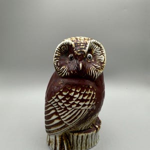 May include: A ceramic owl figurine in shades of brown and white. The owl is perched on a tree stump base. The owl's feathers are detailed with white lines, and it has large, round eyes.