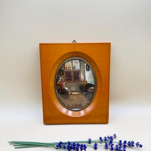 May include: A rectangular orange frame with an oval center opening containing a miniature painting of an interior scene with a person sitting on a chair near a window. The frame has a small metal hanging loop at the top center. A sprig of dried purple lavender lies in front of the frame.