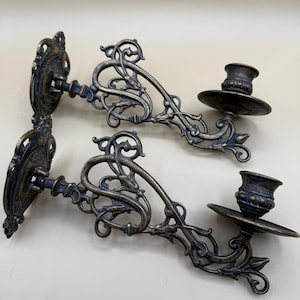 May include: Two antique wall-mounted candle holders. The holders are made of a dark metal with an ornate, swirling design. Each holder has a small, round cup to hold a candle. The design includes floral and scrollwork elements.