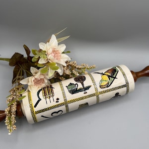 Puede incluir: Un rodillo de amasar vintage con un cuerpo de cerámica crema y mangos de madera. El cuerpo presenta ilustraciones en negro y amarillo de artículos de cocina. Flores decorativas dispuestas cerca del rodillo.