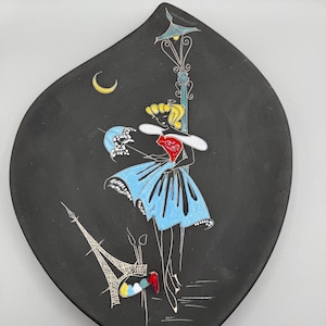 May include: Black decorative plate with a stylized illustration of a woman in a blue dress, a street lamp, the Eiffel Tower, and a crescent moon. The plate has a unique, teardrop shape.