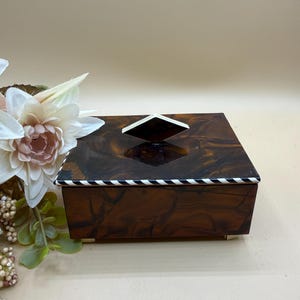 May include: A rectangular box with a glossy brown marbled finish, featuring a black and white striped trim along the lid edge and a diamond-shaped white handle on top. The box is supported by small, light-colored feet at the base.