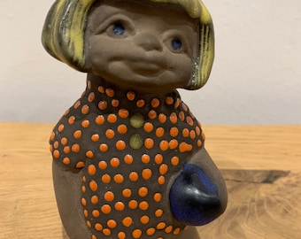 May include: A ceramic figurine of a girl with yellow hair, wearing a brown and orange polka dot dress and holding a blue ball.