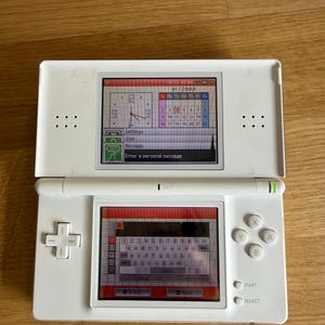 May include: White Nintendo DS handheld game console, open to display two screens. The top screen shows a calendar and clock interface. The bottom screen displays a keyboard for text input. The console has a directional pad, action buttons, and start/select buttons.