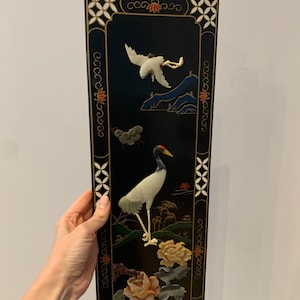 Puede incluir: Un panel de pared lacado en negro con un marco dorado y tallas intrincadas. El panel presenta una grúa de jade blanco y motivos florales en relieve.