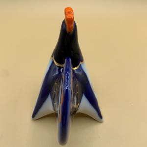 May include: A blue and white porcelain rooster figurine with gold accents. The rooster is standing on its feet with its tail feathers spread out. The rooster's head is tilted back and its beak is open.