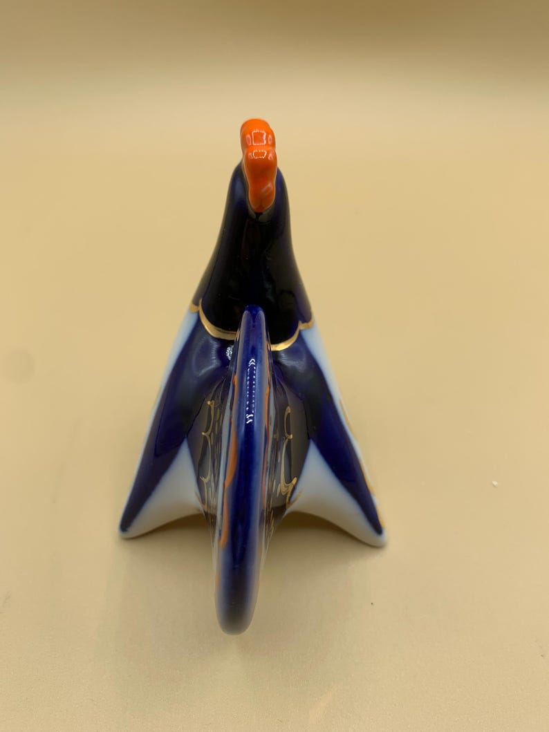 May include: A blue and white porcelain rooster figurine with gold accents. The rooster is standing on its feet with its tail feathers spread out. The rooster's head is tilted back and its beak is open.