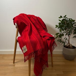 May include: A vibrant red woven throw blanket featuring subtle vertical stripes in darker red and orange tones. The blanket has a fringed edge and a rectangular white fabric label with the text "Tighfield" stitched onto the lower corner.