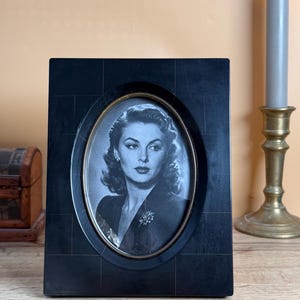May include: A rectangular black picture frame with a grid-like pattern and a central oval opening. The frame holds a black and white portrait of a person with wavy hair wearing a dark blazer and a brooch. The inner edge of the oval is gold-toned.