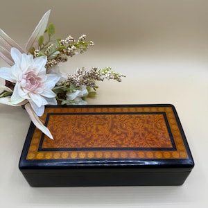 Könnte beinhalten: Eine rechteckige schwarze Box mit dekorativem Deckel. Der Deckel hat ein braunes Blumenmuster, das von einem schwarzen Rand mit goldenen Akzenten umrahmt wird. Im Hintergrund befindet sich ein weiß-rosa Blumenarrangement.
