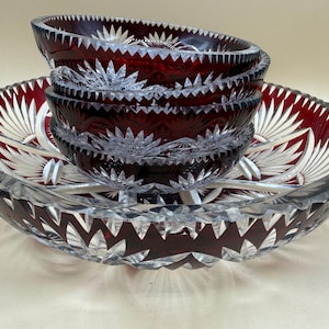 Op de afbeelding: Een set dieprode en heldere geslepen glazen schalen met een sterpatroon. De collectie bevat één grote ondiepe schaal en drie kleinere gestapelde schalen, allemaal met ingewikkelde geometrische facetten en een gekartelde rand.