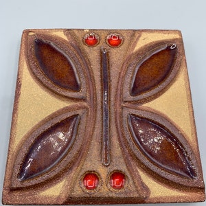 Könnte beinhalten: Eine quadratische Keramikfliese mit brauner und beigefarbener Glasur. Die Fliese zeigt ein erhabenes Design eines Schmetterlings mit roten Glasakzenten.