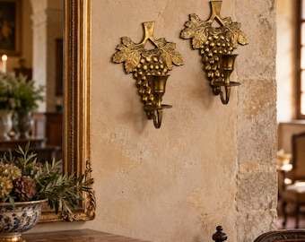 May include: Two gold-toned metal wall sconces shaped like grape clusters with leaves. Each sconce features a central candle holder cup attached to the bottom of the textured grape design, designed for mounting on a flat vertical surface.