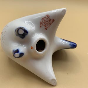 May include: White ceramic figurine of a stylized cat with blue and red accents. The figurine has a hole in the top and the text "USSR MADE IN USSR" on the back.