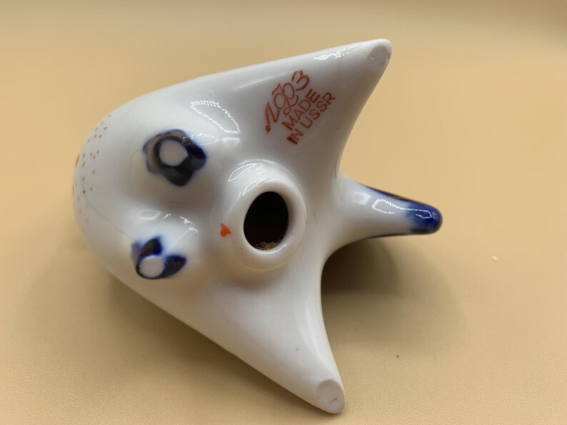 May include: White ceramic figurine of a stylized cat with blue and red accents. The figurine has a hole in the top and the text "USSR MADE IN USSR" on the back.