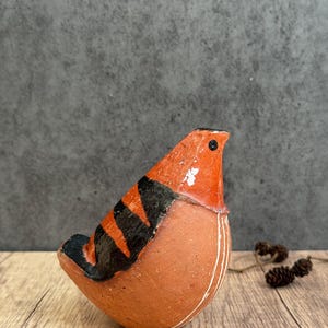 Może przedstawiać: Mała, zaokrąglona figurka ptaka wykonana z gliny w kolorze terakoty. Ptak ma błyszczącą pomarańczową głowę i grzbiet z wyraźnymi, czarnymi ukośnymi paskami na skrzydłach i ogonie. Białe linie zdobią pierś oraz bok figurki ptaka.
