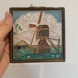 Könnte beinhalten: Eine Keramikfliese mit einem gerahmten Bild einer Windmühle in einer Landschaft. Die Windmühle ist braun und hat ein weißes Segel. Der Hintergrund ist blau und weiß mit einem bewölkten Himmel.