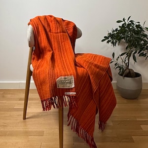 Puede incluir: Una manta tejida de color naranja vibrante con un patrón de rayas texturizado y bordes con flecos. Una etiqueta de tela blanca rectangular está cosida en la parte delantera, con el texto Tidstrand y otros detalles impresos.